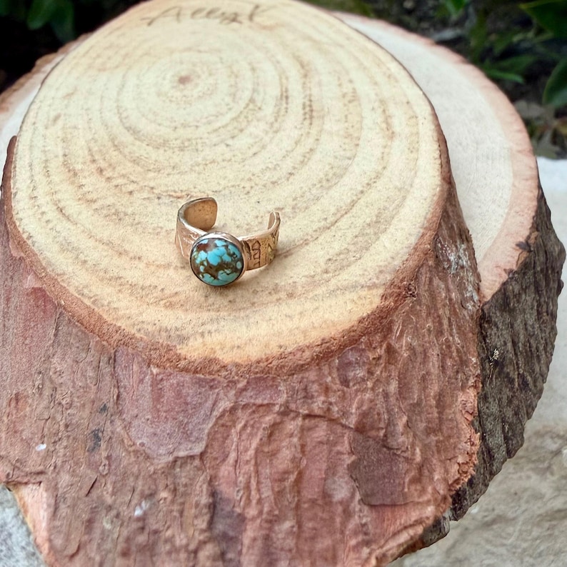 May include: A gold-colored ring with a round, turquoise stone. The ring is set on a wooden surface. The ring's band has etched details. The turquoise stone has a mix of blue and brown colors.