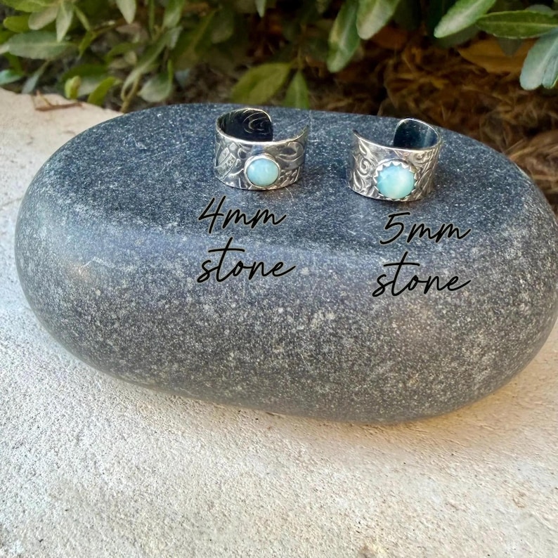 May include: Two silver rings with textured bands and light blue circular stones. One ring features a 4mm stone, and the other features a 5mm stone. The rings are displayed on a smooth, dark gray stone surface with text labels indicating the stone sizes.