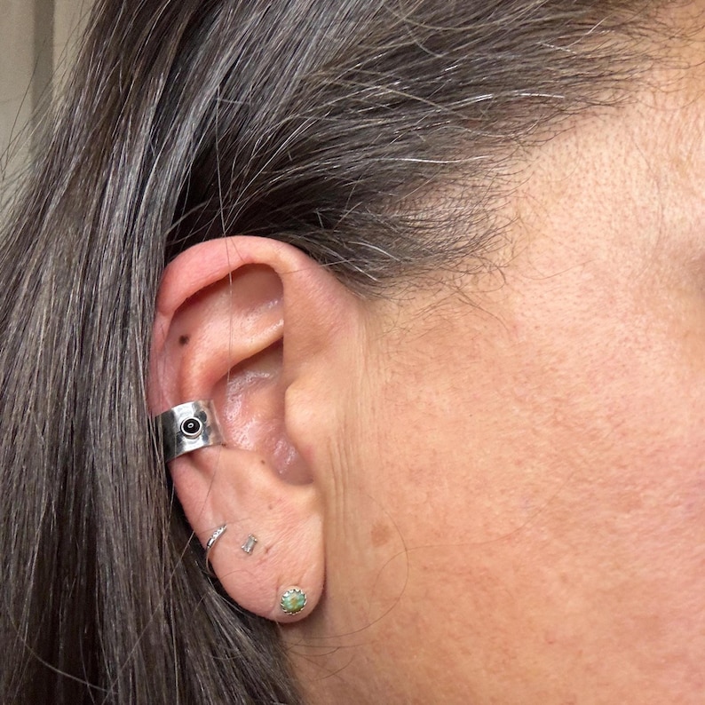 May include: A silver ear cuff with a black stone is worn on the ear. The ear also has a small silver hoop earring, a small silver stud earring, and a small green stud earring.