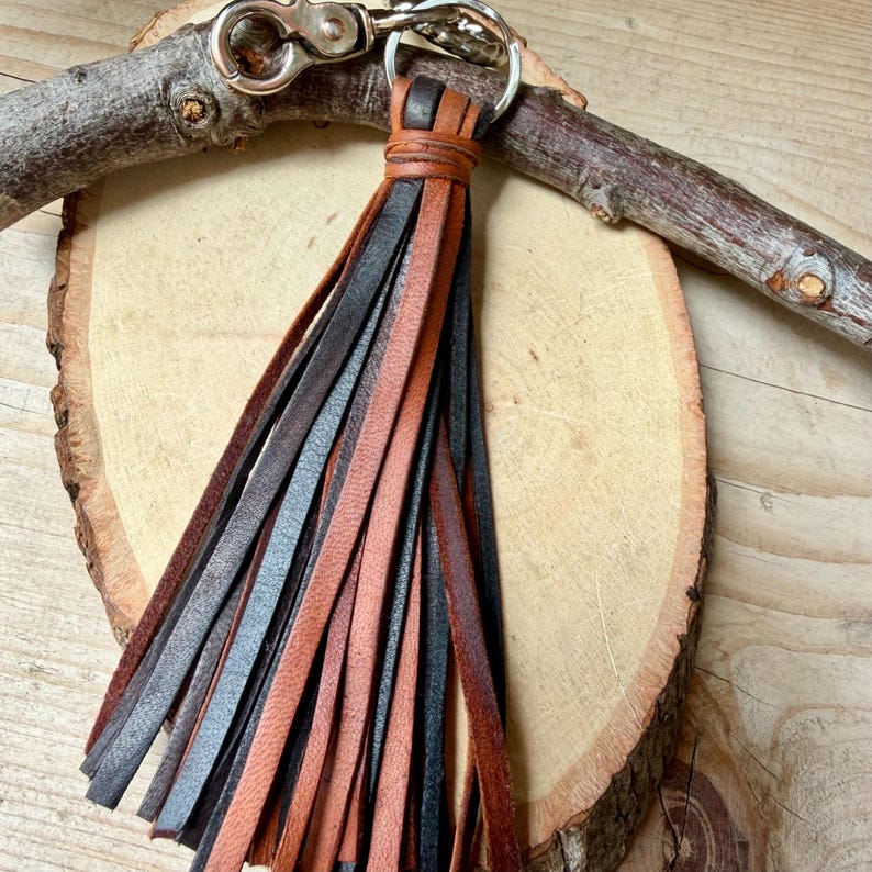 May include: A leather tassel keychain with a silver clasp. The tassel is made of strips of brown, black, and tan leather. The keychain is displayed on a wooden slice and a small branch.