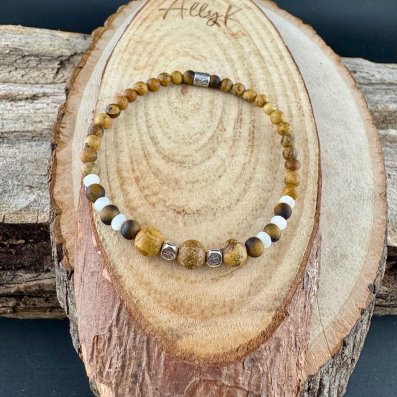 May include: A beaded bracelet featuring a mix of brown, tan, and white beads. The bracelet includes larger, textured beads and smaller, round beads. The clasp is silver-toned. The bracelet is displayed on a wooden surface.