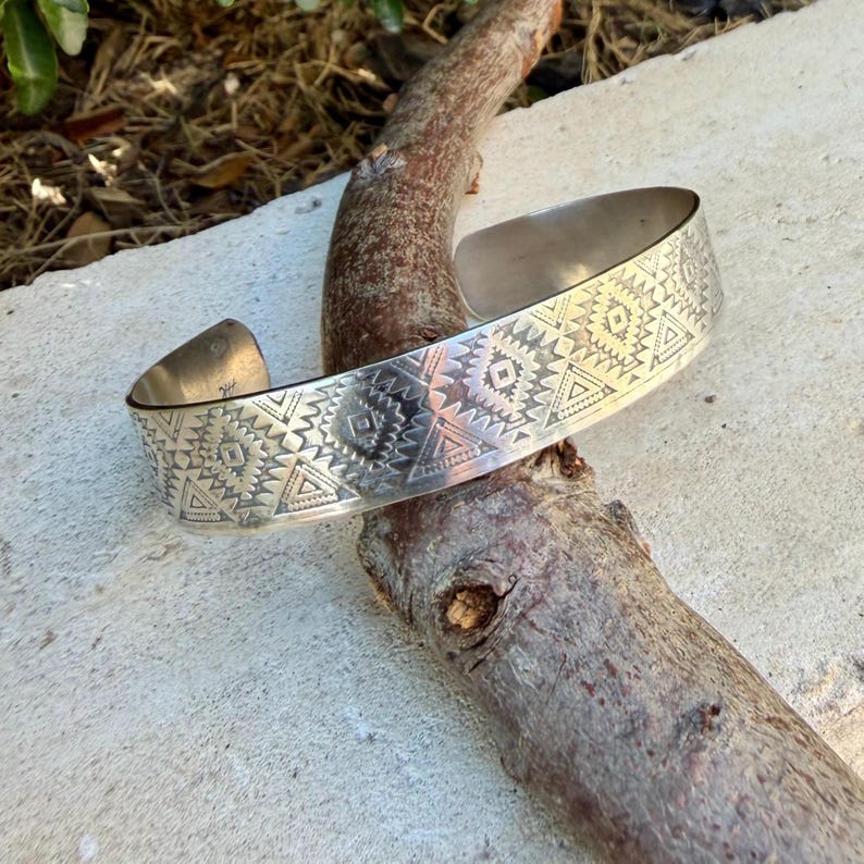 May include: A silver cuff bracelet featuring a repeating geometric pattern of triangles, diamonds, and linear etchings across the outer surface. The metal band has an open back design and a polished finish, showcasing intricate stamped detailing throughout.