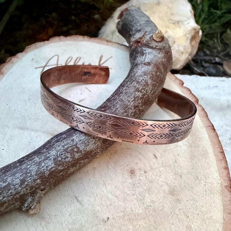 May include: A copper cuff bracelet featuring a repeating geometric pattern of etched diamonds, chevrons, and horizontal lines. The metal has a warm, reddish-brown metallic finish with darkened recessed areas that highlight the intricate stamped design.