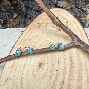 May include: Four pairs of turquoise stud earrings with silver settings are displayed on a wooden branch. The earrings have a round, polished turquoise stone. The branch rests on a wooden surface with the word "Alex" written on it.