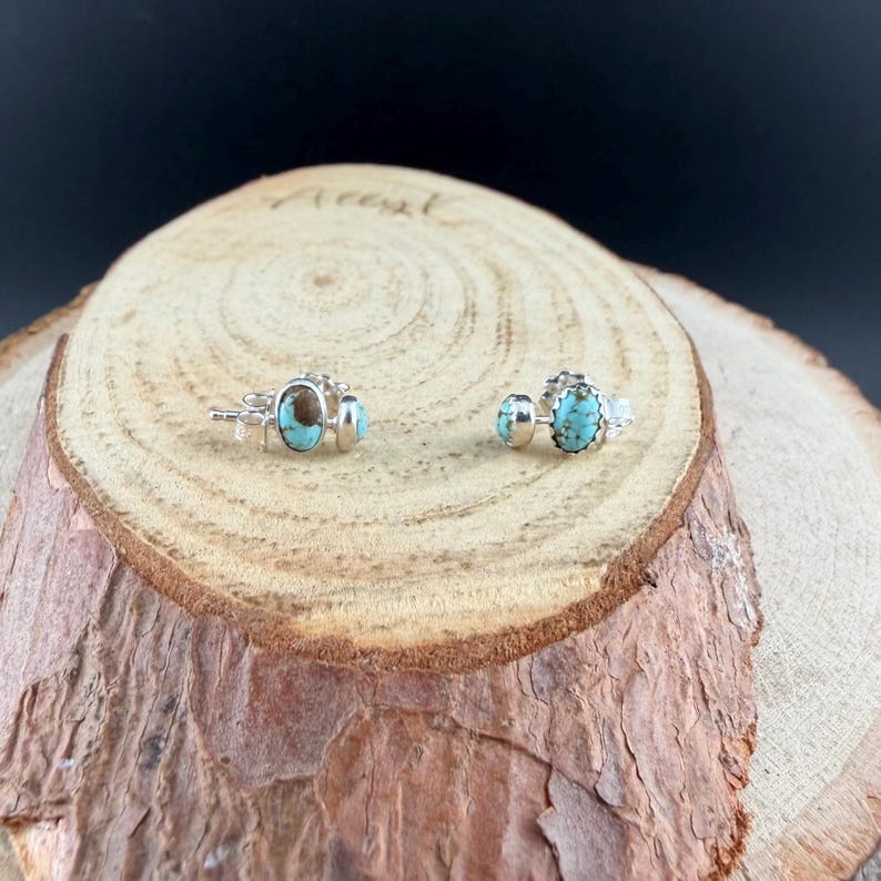 May include: A pair of silver stud earrings with oval turquoise and brown stones. The earrings are displayed on a wooden surface with a dark background. The earrings have a detailed, decorative setting.