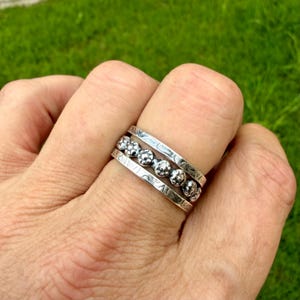 May include: A silver ring with a textured design and a row of small, round embellishments. The ring has a detailed pattern on the band and is worn on a finger. The ring is made of a shiny metal.