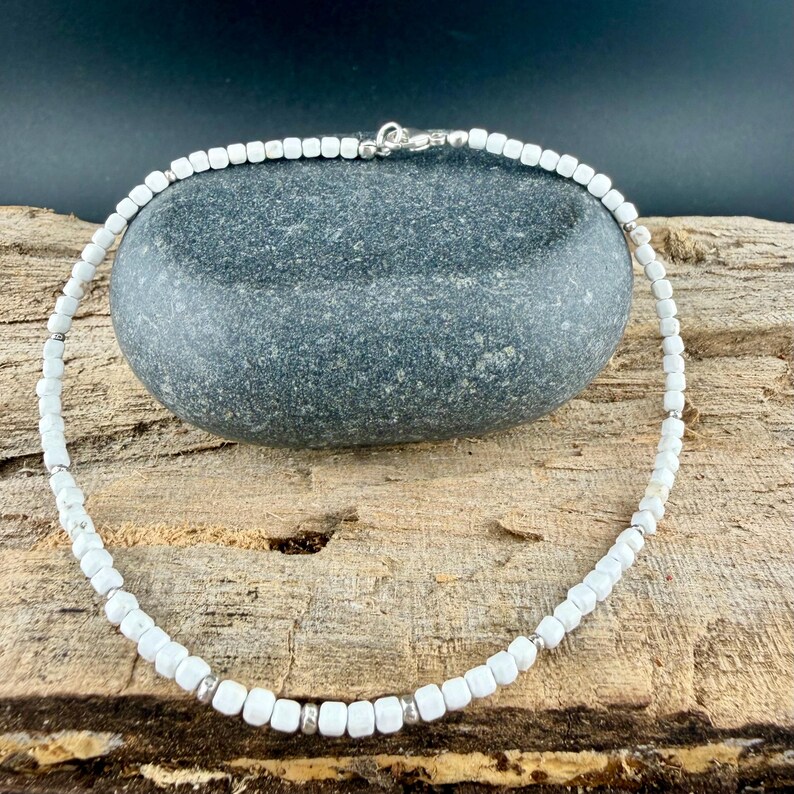 May include: White beaded necklace with silver accents. The necklace features small, square white beads strung together with silver-colored metal spacers and a clasp. The necklace is displayed on a gray stone against a wooden background.