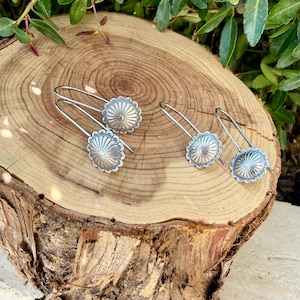 May include: Two pairs of silver-toned earrings featuring a circular, sunburst-patterned design with radiating lines. Each earring has a curved wire hook for attachment, showcasing a metallic finish with a textured, embossed radial detail.