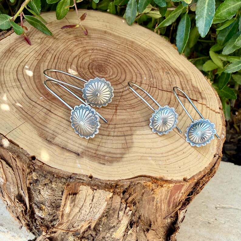 May include: Two pairs of silver-toned earrings featuring a circular, sunburst-patterned design with radiating lines. Each earring has a curved wire hook for attachment, showcasing a metallic finish with a textured, embossed radial detail.