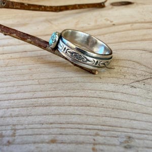 Puede incluir: Un anillo de plata con una banda pulida que presenta un patr&oacute;n floral grabado y una peque&ntilde;a piedra turquesa redonda engastada en un bisel dentado. El anillo est&aacute; equilibrado sobre una rama de madera fina y marr&oacute;n.