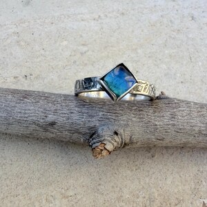 May include: A silver ring with a square, blue and teal-colored stone. The band has a textured design. The ring is displayed on a wooden branch against a neutral background. The ring's design is geometric and modern.