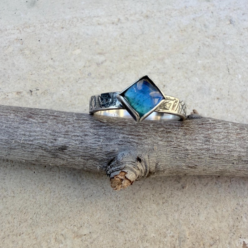 May include: A silver ring with a square, blue and teal-colored stone. The band has a textured design. The ring is displayed on a wooden branch against a neutral background. The ring's design is geometric and modern.