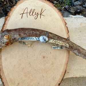 May include: A silver cuff bracelet with a white and grey stone in the centre. The bracelet has decorative etched designs. The word "Allyk" is written in cursive on a wooden background.
