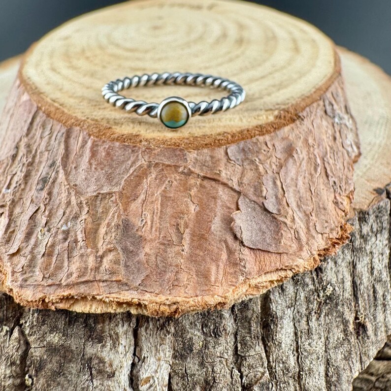 May include: A silver-toned ring with a twisted band design. The ring features a small, round, light brown and turquoise stone at the center. The ring is displayed on a textured, brown wooden surface.