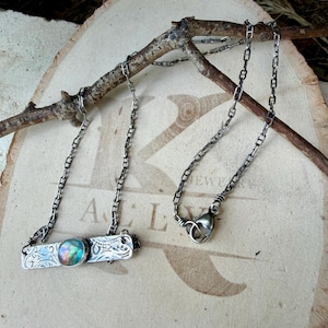 May include: A silver-toned necklace featuring a rectangular metal bar pendant with an embossed floral pattern and a central round iridescent cabochon stone. The pendant is attached to a delicate link chain with a lobster claw clasp.