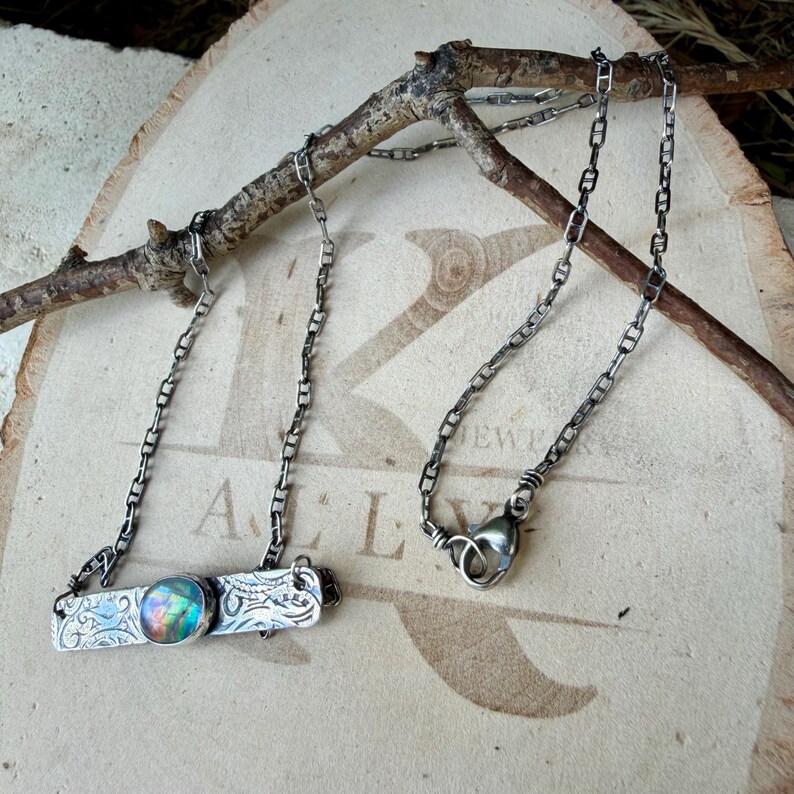 May include: A silver-toned necklace featuring a rectangular metal bar pendant with an embossed floral pattern and a central round iridescent cabochon stone. The pendant is attached to a delicate link chain with a lobster claw clasp.