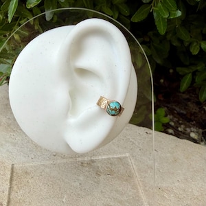 May include: A gold-colored ear cuff with a round, turquoise-colored stone. The cuff is displayed on a white ear-shaped form. The cuff has a textured band with a decorative pattern. The turquoise stone is set in a bezel setting.