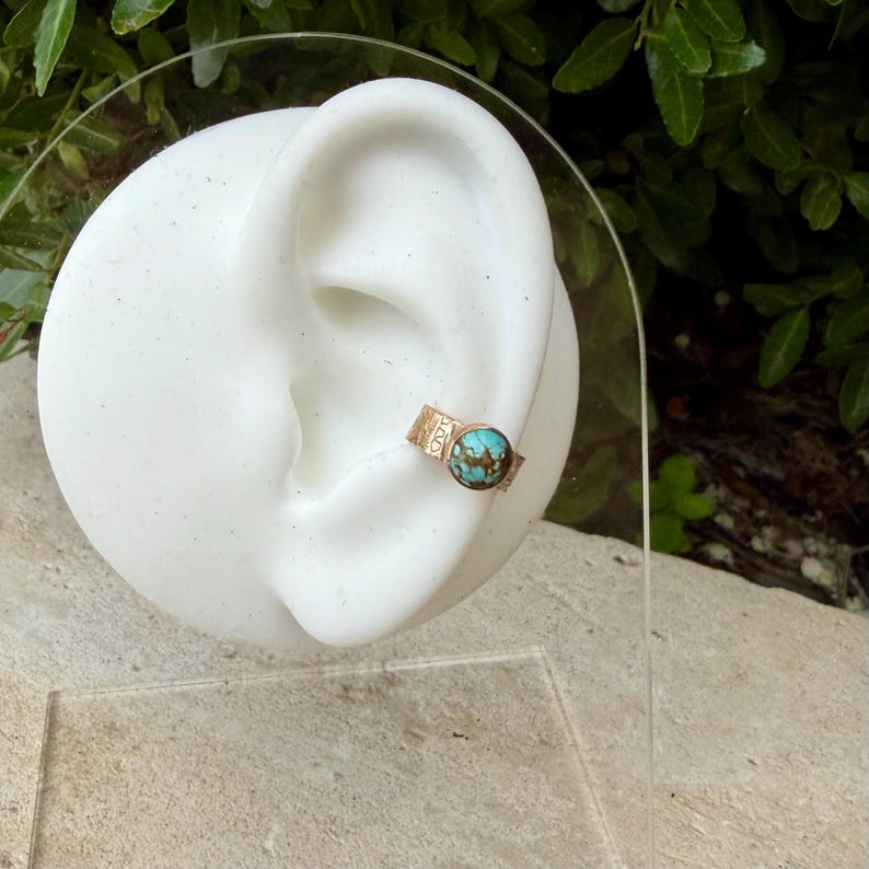 May include: A gold-colored ear cuff with a round, turquoise-colored stone. The cuff is displayed on a white ear-shaped form. The cuff has a textured band with a decorative pattern. The turquoise stone is set in a bezel setting.