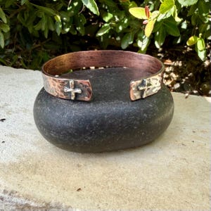 May include: A copper cuff bracelet featuring a hammered texture and a polished finish. The open-ended band displays two silver-toned cross emblems positioned near the terminals, providing a distinct contrast against the warm metallic surface.