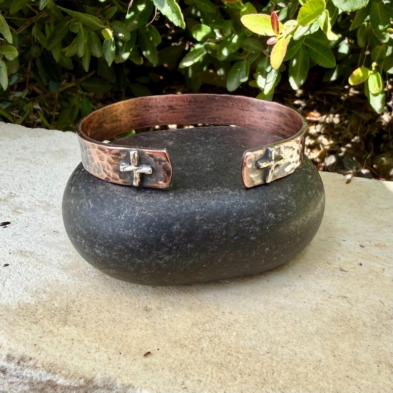 May include: A copper cuff bracelet featuring a hammered texture and a polished finish. The open-ended band displays two silver-toned cross emblems positioned near the terminals, providing a distinct contrast against the warm metallic surface.