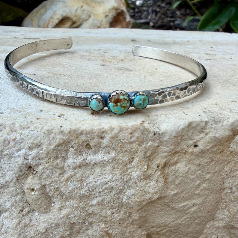 May include: A silver cuff bracelet with a hammered texture and three turquoise stones. The bracelet is open-ended and has a simple, elegant design. The turquoise stones are round and set in a silver bezel.