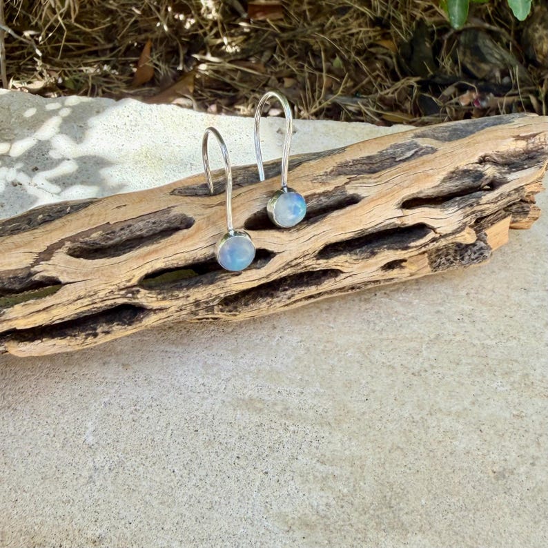 May include: A pair of silver drop earrings featuring round, iridescent moonstone cabochons. Each earring has a curved silver wire hook that suspends the circular stone setting, showcasing a soft blue and white glow within the polished gems.