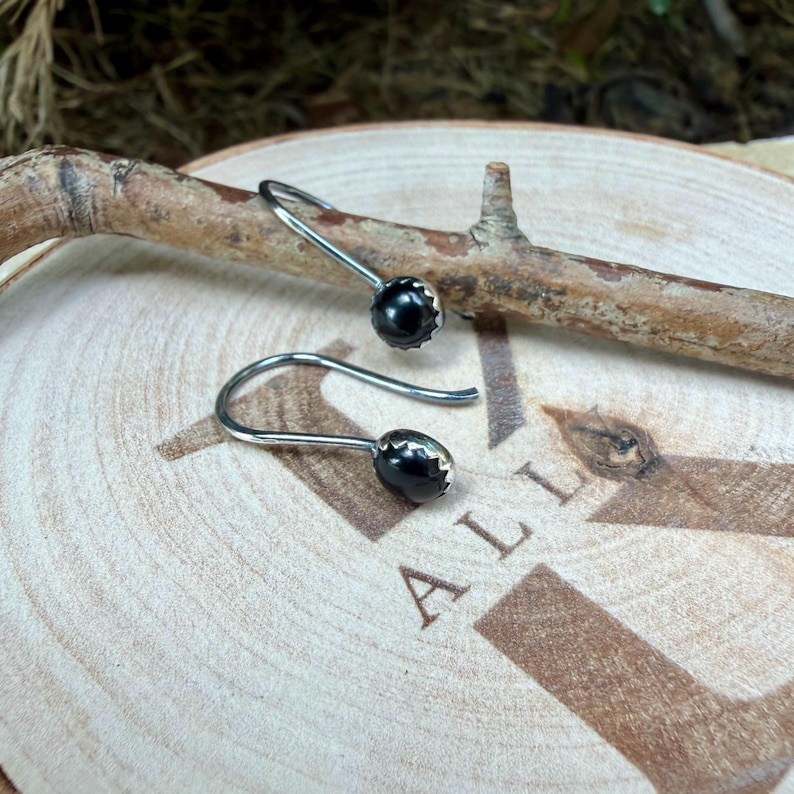 May include: A pair of silver hook earrings featuring small, round, polished black stones. Each earring has a curved metal wire that leads to a smooth, dark spherical bead set in a simple silver mounting, creating a clean and reflective appearance.