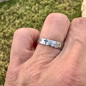 May include: A hammered silver ring with a cross design. The ring has a textured surface and a simple, elegant style. The cross is centered on the band. The ring is shown on a finger.