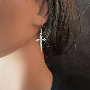 May include: A silver-toned drop earring featuring a slender, pointed dagger-shaped pendant with ornate, filigree-style detailing at the hilt. The earring hangs from a simple, thin wire hook, showcasing a metallic finish and a sharp, tapered design.