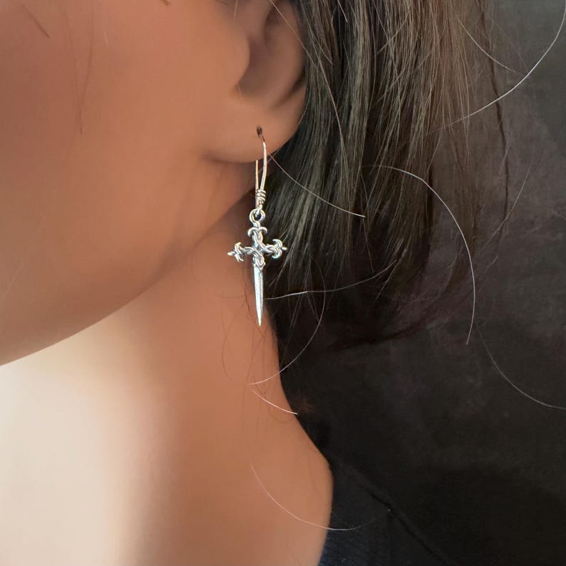 May include: A silver-toned drop earring featuring a slender, pointed dagger-shaped pendant with ornate, filigree-style detailing at the hilt. The earring hangs from a simple, thin wire hook, showcasing a metallic finish and a sharp, tapered design.