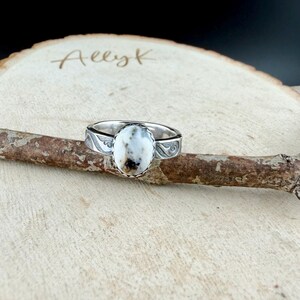 May include: A silver ring with an oval, white stone with brown and black inclusions. The ring band features an engraved design. The ring is displayed on a wooden branch, with the name "Aeezk" written on the wooden surface in the background.