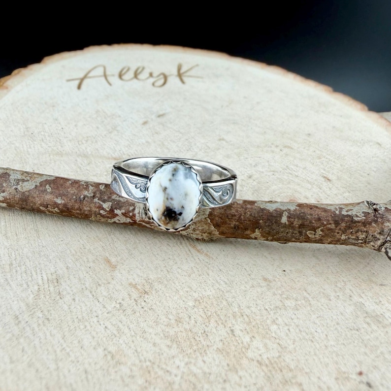 May include: A silver ring with an oval, white stone with brown and black inclusions. The ring band features an engraved design. The ring is displayed on a wooden branch, with the name "Aeezk" written on the wooden surface in the background.