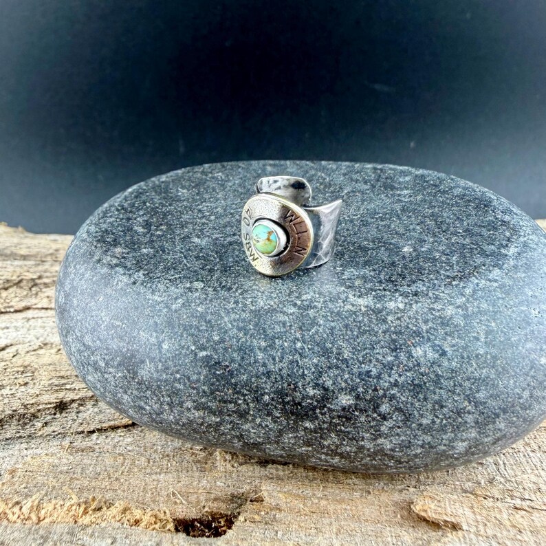 May include: A silver ring featuring a turquoise stone set within a brass casing. The ring's band wraps around the finger, creating a distinctive design. The ring is presented on a grey stone surface.