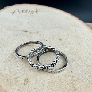 May include: Three silver rings displayed on a wooden surface. One ring has a beaded design, another has etched patterns, and the third is a simple band. The rings are arranged in a layered composition, showcasing their individual designs.