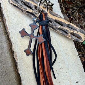 Op de afbeelding: Een sleutelhanger met een bruine leren kruishanger en een meerstrengige kwast van zwarte en lichtbruine leren stroken. De leren elementen zijn bevestigd aan een zilverkleurige metalen ring, wat zorgt voor een decoratief accessoire met gelaagd design.
