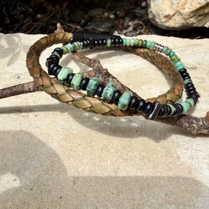May include: Two bracelets with a natural, earthy aesthetic. One is braided leather, the other features turquoise and black beads. The bracelets are displayed on a small branch, showcasing their textures and colors.
