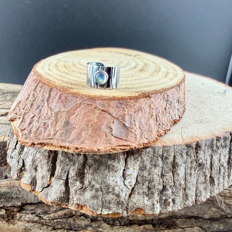 K&ouml;nnte beinhalten: Ein breiter Silberring mit strukturiertem Band und einem zentralen, schillernden, oval geformten Stein. Der Ring wird auf einer rustikalen Holzoberfl&auml;che pr&auml;sentiert, die sein Design hervorhebt. Das Design des Rings ist modern und auff&auml;llig.