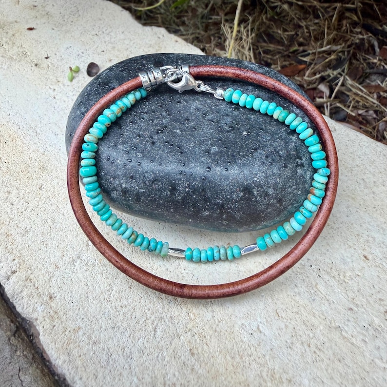May include: A double-strand bracelet featuring one strand of small, round, turquoise-colored beads with silver accents and one strand of smooth, brown leather cord. The bracelet is secured with a silver-toned lobster claw clasp and metal end caps.