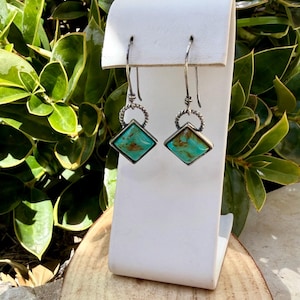 May include: A pair of silver-toned dangle earrings. Each earring features a square, turquoise stone with brown veining, set in a silver frame. The stones are suspended from a twisted silver ring and a simple hook.
