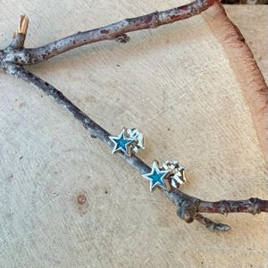 May include: A pair of star-shaped stud earrings with a silver finish and turquoise enamel centers. The earrings are displayed on a wooden branch against a light wood background. The earrings are a celestial-themed accessory.