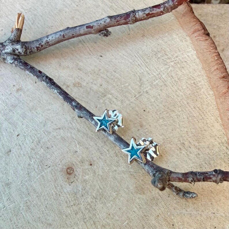 May include: A pair of star-shaped stud earrings with a silver finish and turquoise enamel centers. The earrings are displayed on a wooden branch against a light wood background. The earrings are a celestial-themed accessory.