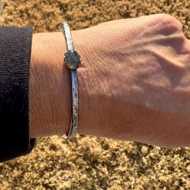 May include: A silver-toned cuff bracelet with a textured surface and a central, oval-shaped, gray gemstone. The bracelet is worn on a wrist, showcasing its design and craftsmanship. The cuff is approximately 2.5 inches in diameter.