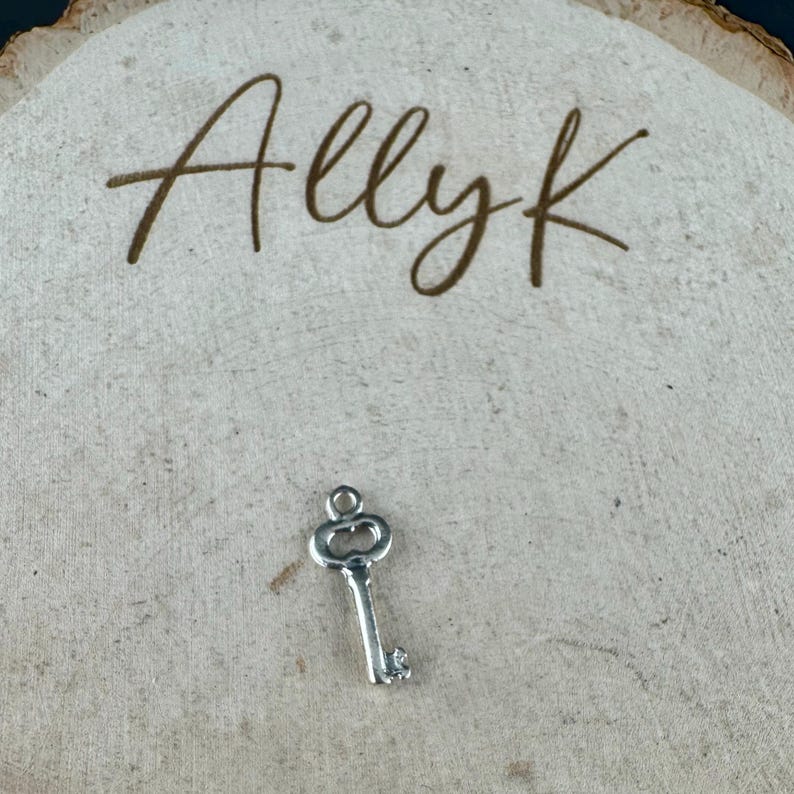 May include: A small, silver-colored key charm with a decorative top and a detailed key bit. The charm is set against a light-colored, textured wooden background. The word "Allyk" is written in cursive script above the key.