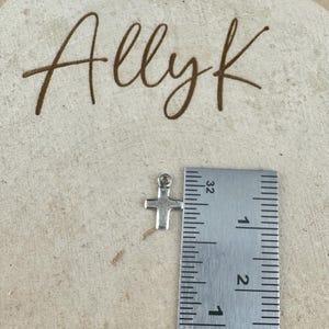 May include: A small, silver-colored cross charm with a loop at the top, next to a ruler. The ruler shows measurements in inches. The word "Allyk" is written in brown script in the background.