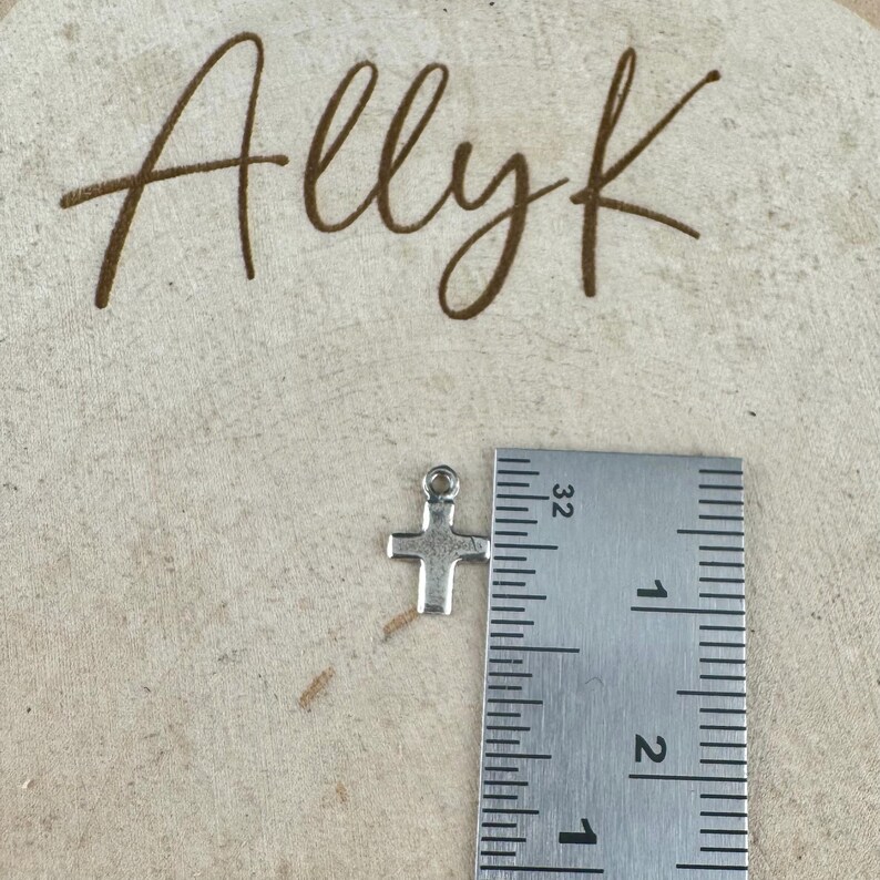 May include: A small, silver-colored cross charm with a loop at the top, next to a ruler. The ruler shows measurements in inches. The word "Allyk" is written in brown script in the background.