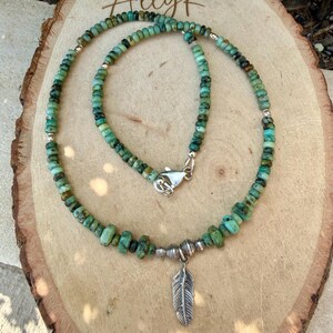 May include: A turquoise beaded necklace with a silver feather pendant. The necklace features small, irregular turquoise beads and silver accents. The feather pendant hangs from a silver clasp.