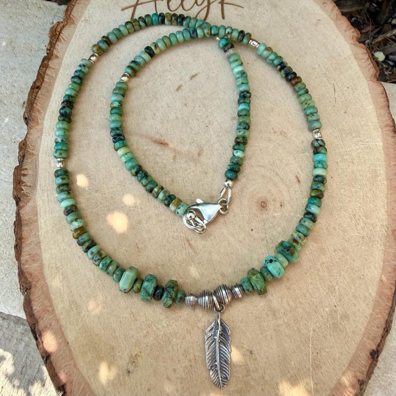 May include: A turquoise beaded necklace with a silver feather pendant. The necklace features small, irregular turquoise beads and silver accents. The feather pendant hangs from a silver clasp.