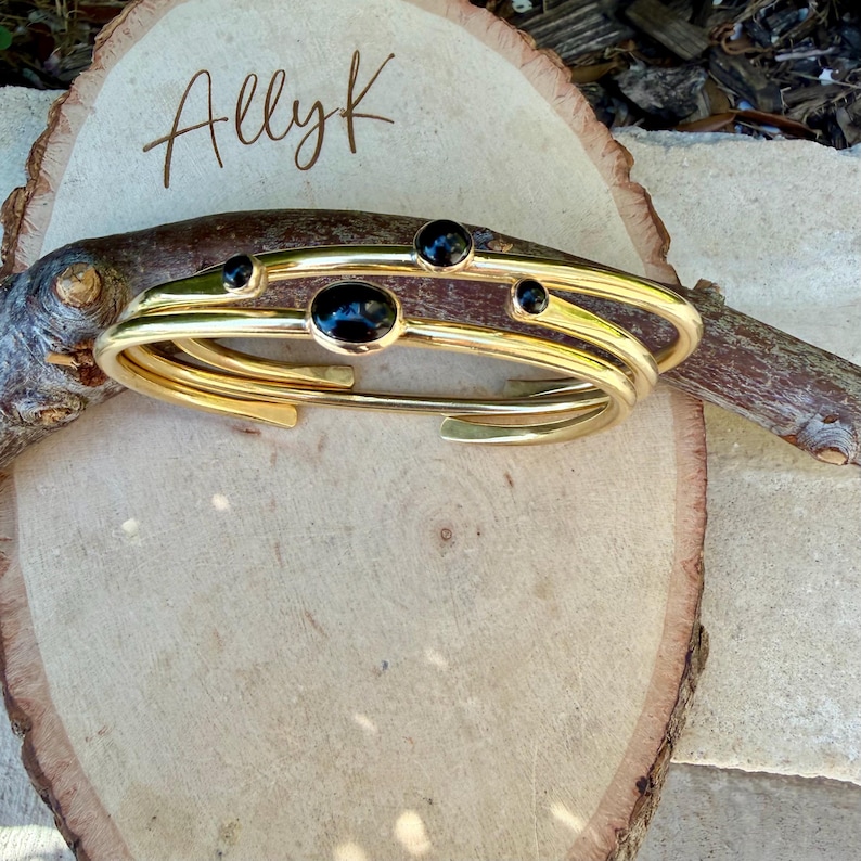 May include: Gold-toned cuff bracelet with four black oval and round stones. The bracelet has a layered design and is displayed on a wooden surface. The word "Allyk" is written in cursive on the wood.