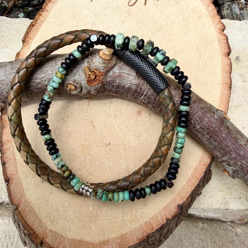 May include: Two bracelets are displayed. One is a braided brown leather bracelet. The other is a beaded bracelet with black and turquoise beads. The bracelets are resting on a wooden surface with a natural wood grain.