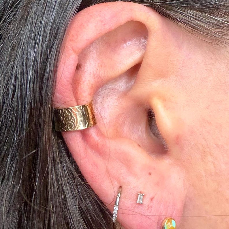 May include: Gold-colored ear cuff with an engraved floral design. The cuff is worn on the upper ear. Additional earrings include a small hoop with clear stones and a small rectangular stud.